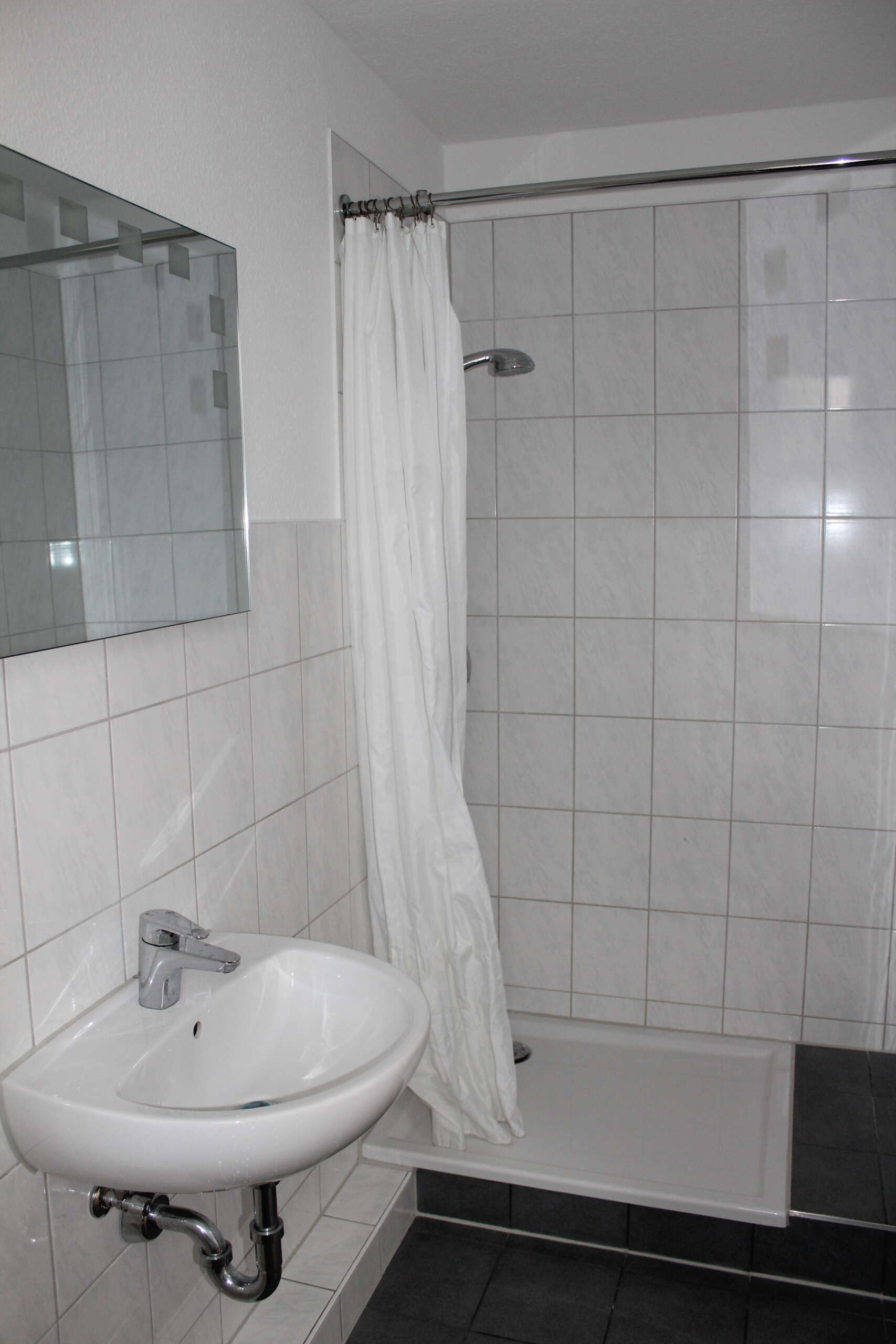 Einfache, geflieste Dusche mit Duschvorhang und Waschbecken im Badezimmer.