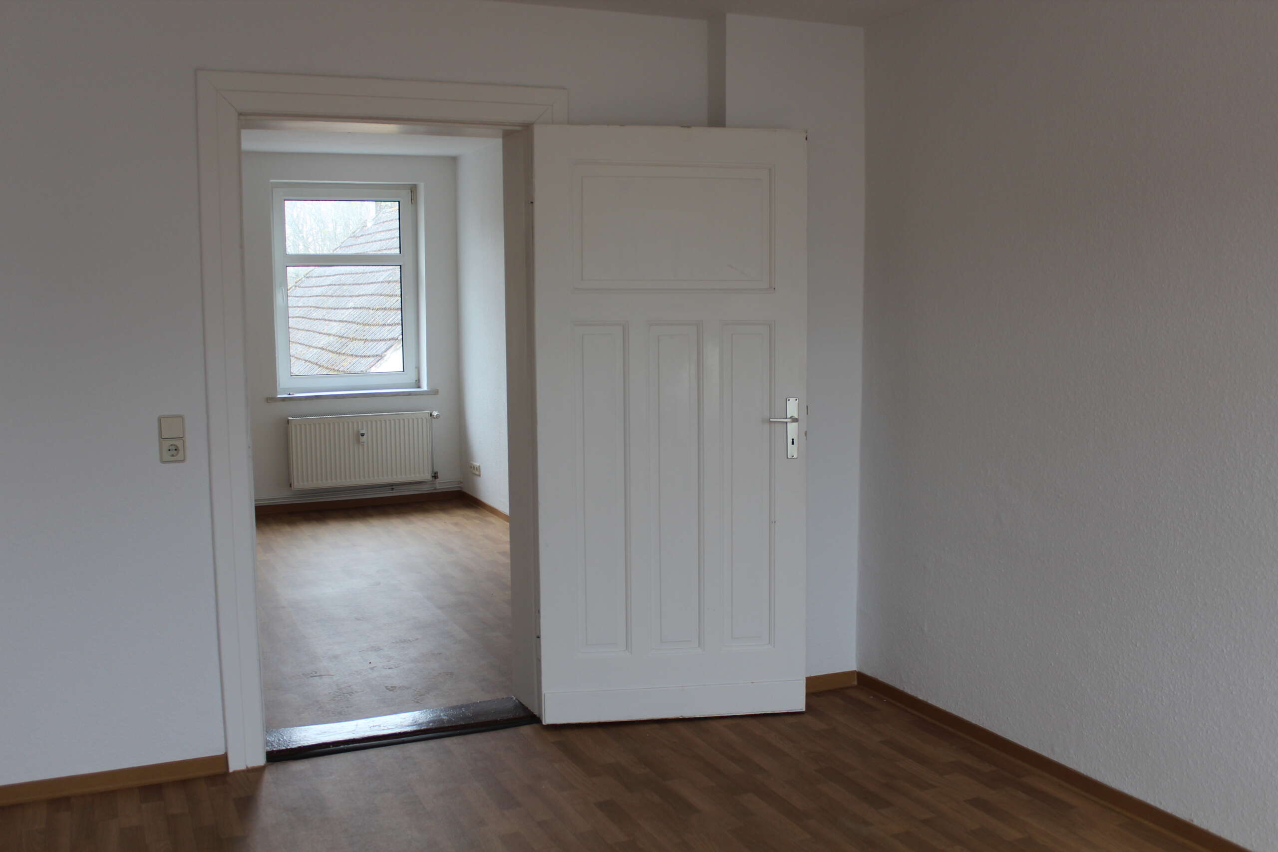 Wohnung mit offenem Türdurchgang, Parkettboden und Fenster, sichtbarer Heizkörper unter dem Fenster.