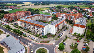 Luftaufnahme eines Wohnkomplexes mit moderner Architektur, umliegenden Feldern und einem Kreisverkehr im Vordergrund.