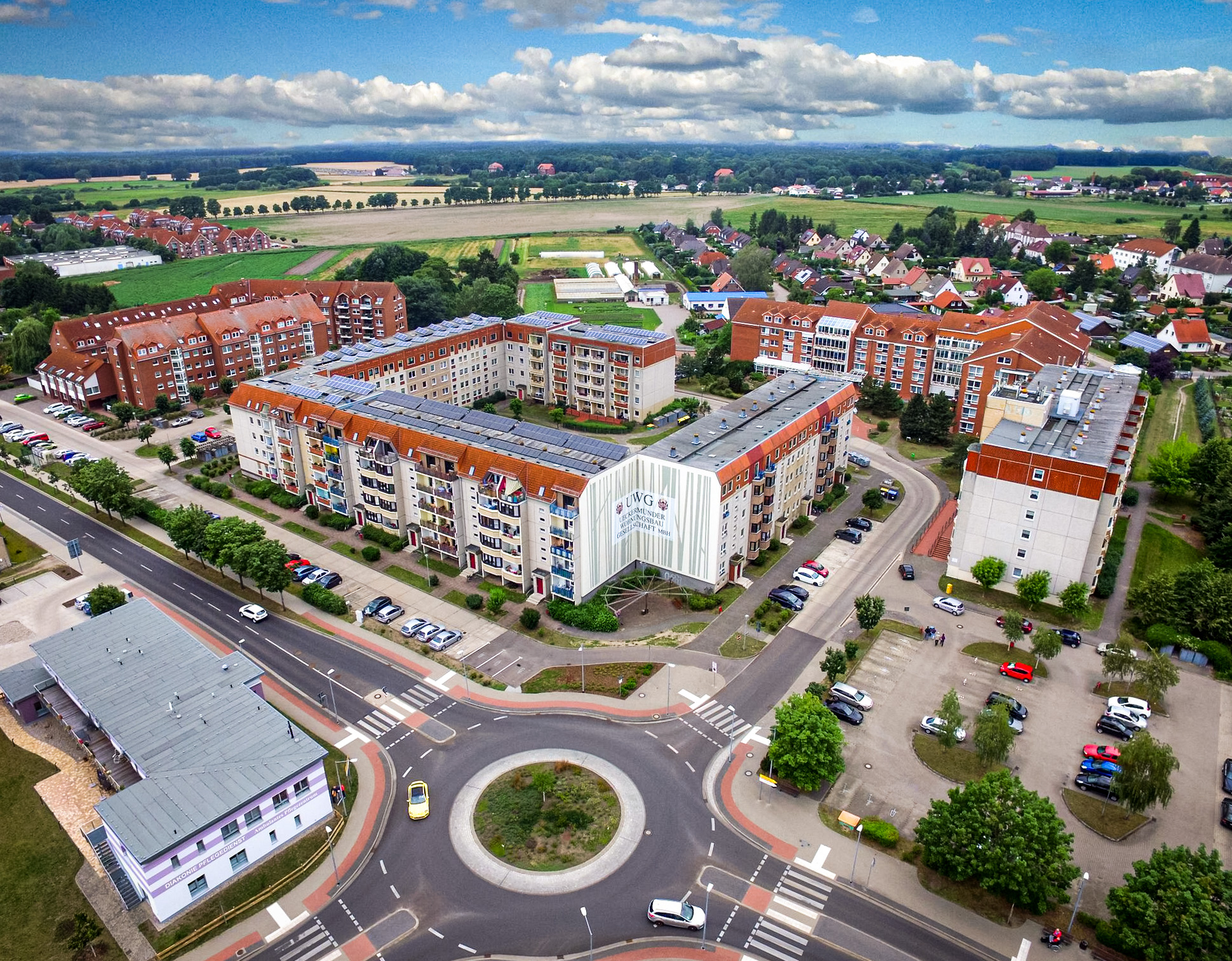 Luftaufnahme eines Wohnkomplexes mit moderner Architektur, umliegenden Feldern und einem Kreisverkehr im Vordergrund.
