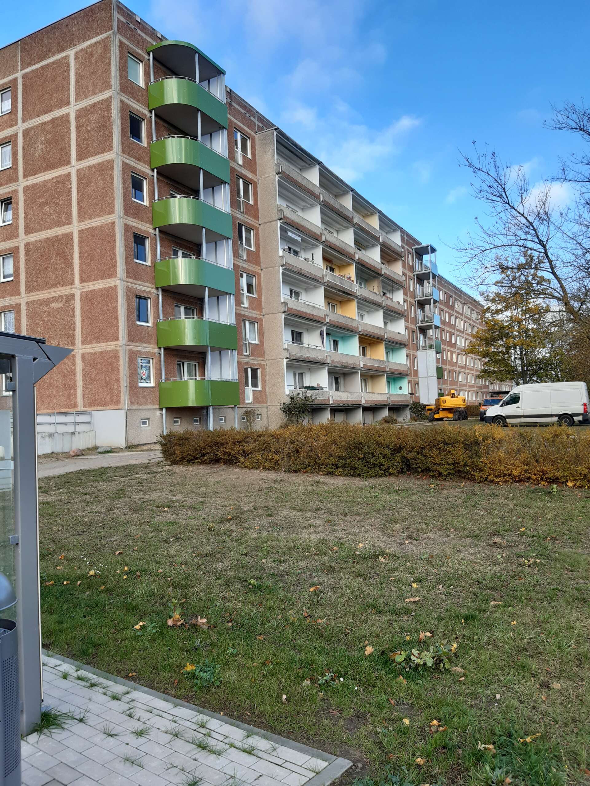 Sechsstöckiges Wohngebäude mit grünen Balkonen, blauer Himmel und herbstliche Bäume im Hintergrund.