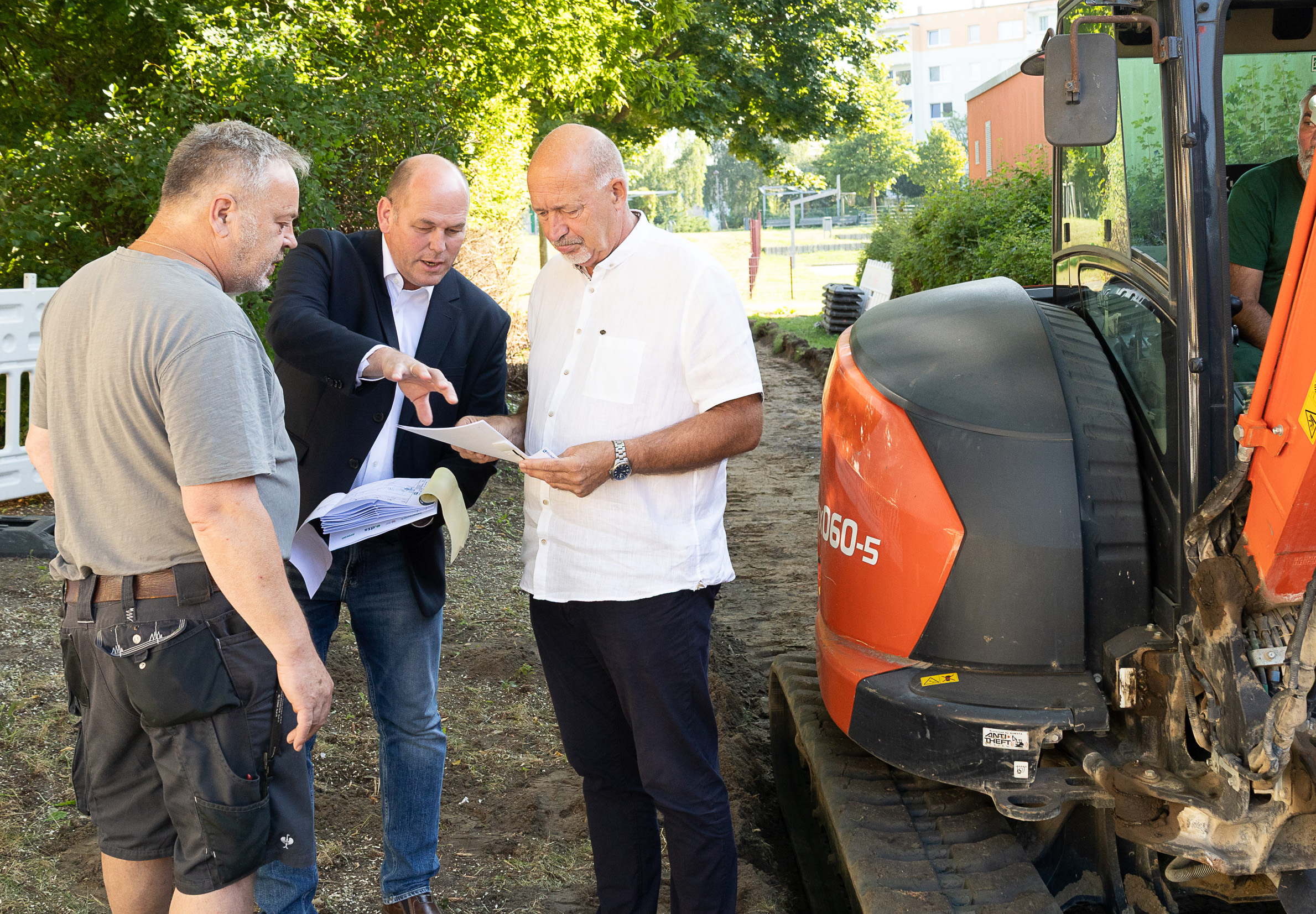 Männer besprechen Baupläne neben einem Bagger auf einer Baustelle im Freien.