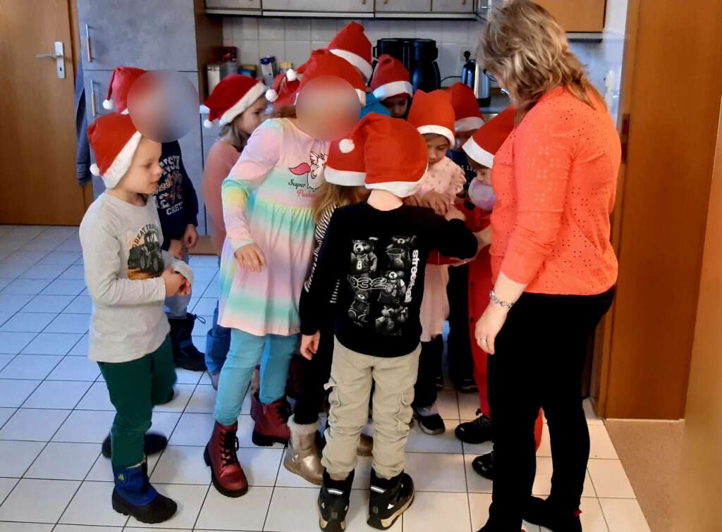 Kinder in Weihnachtsmannmützen erhalten Geschenke in einer Küche. Eine Frau leitet die Gruppe.
