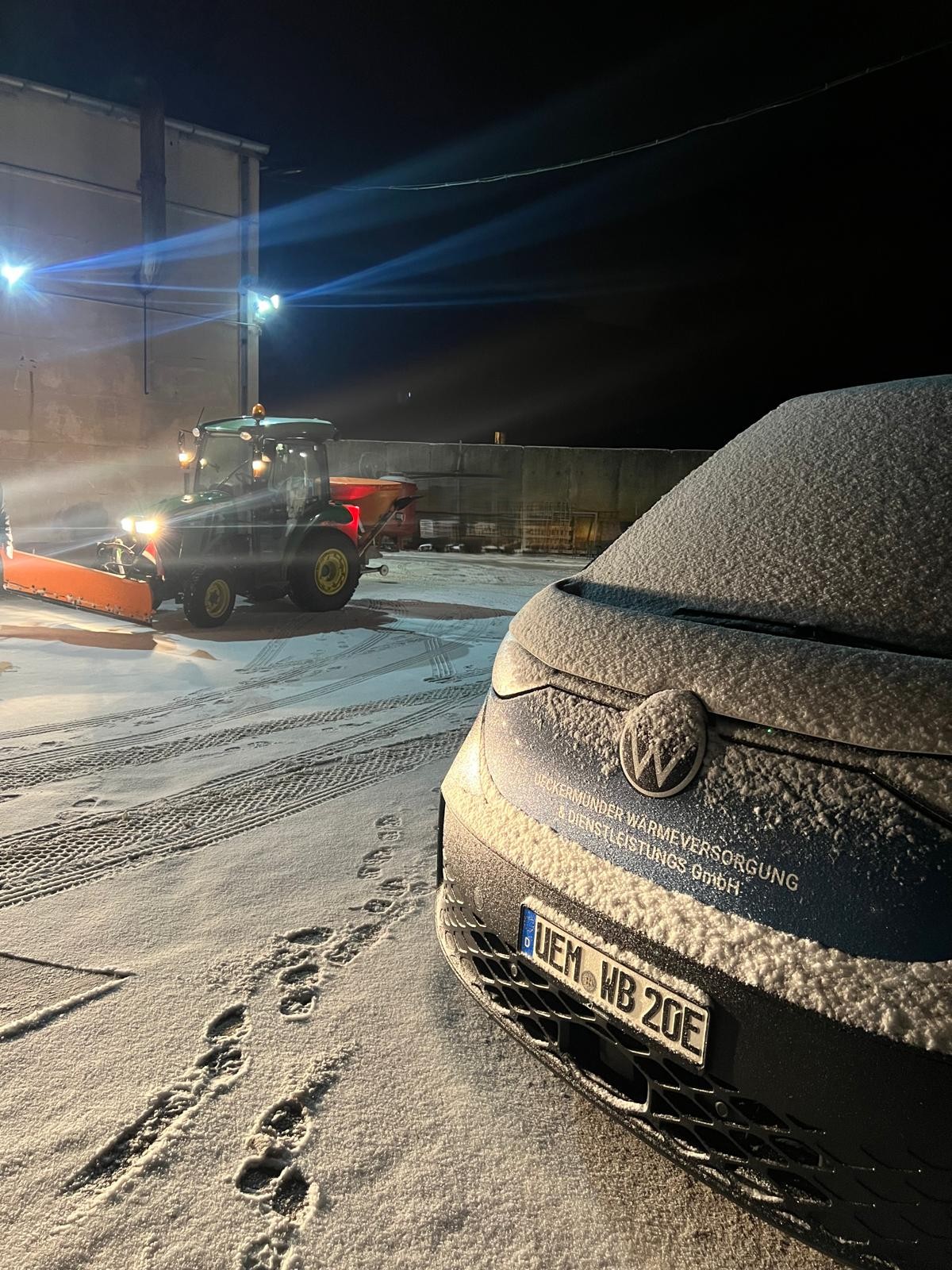 Schneebedecktes Auto und Traktor mit Schneepflug bei Nacht auf verschneitem Gelände, Lichter beleuchten die Szene.