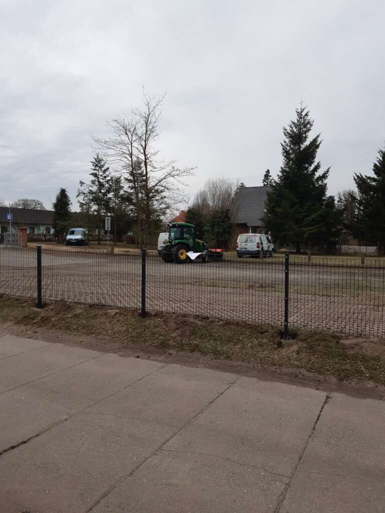 Traktor auf einer Straße vor einem Haus, umgeben von Bäumen, mit bewölktem Himmel im Hintergrund.