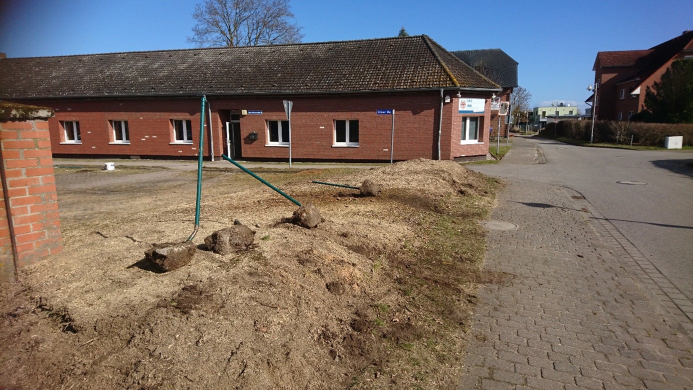 Ziegelgebäude an einer Ecke, Gehweg mit Schildern und Erdarbeiten im Vordergrund unter blauem Himmel.