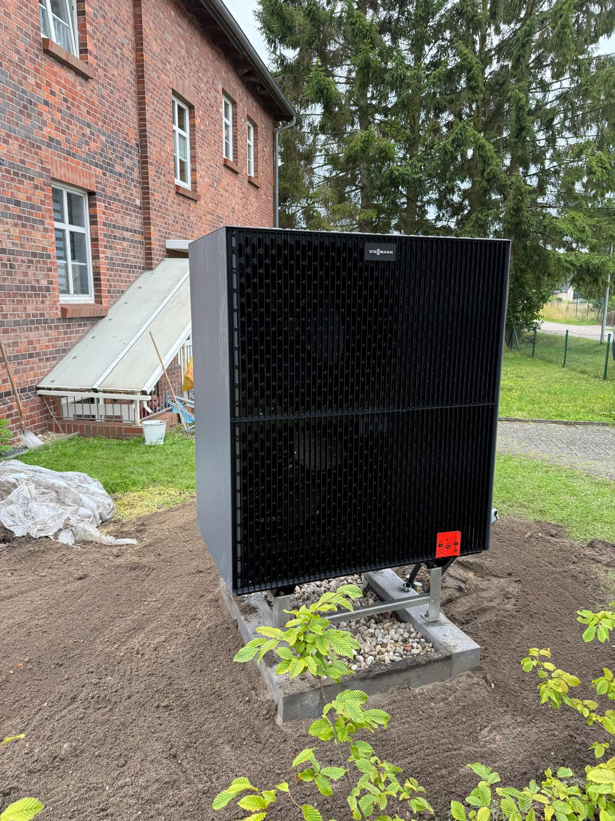 Viessmann Wärmepumpe im Garten neben Backsteingebäude, umgeben von Bäumen und Sträuchern.