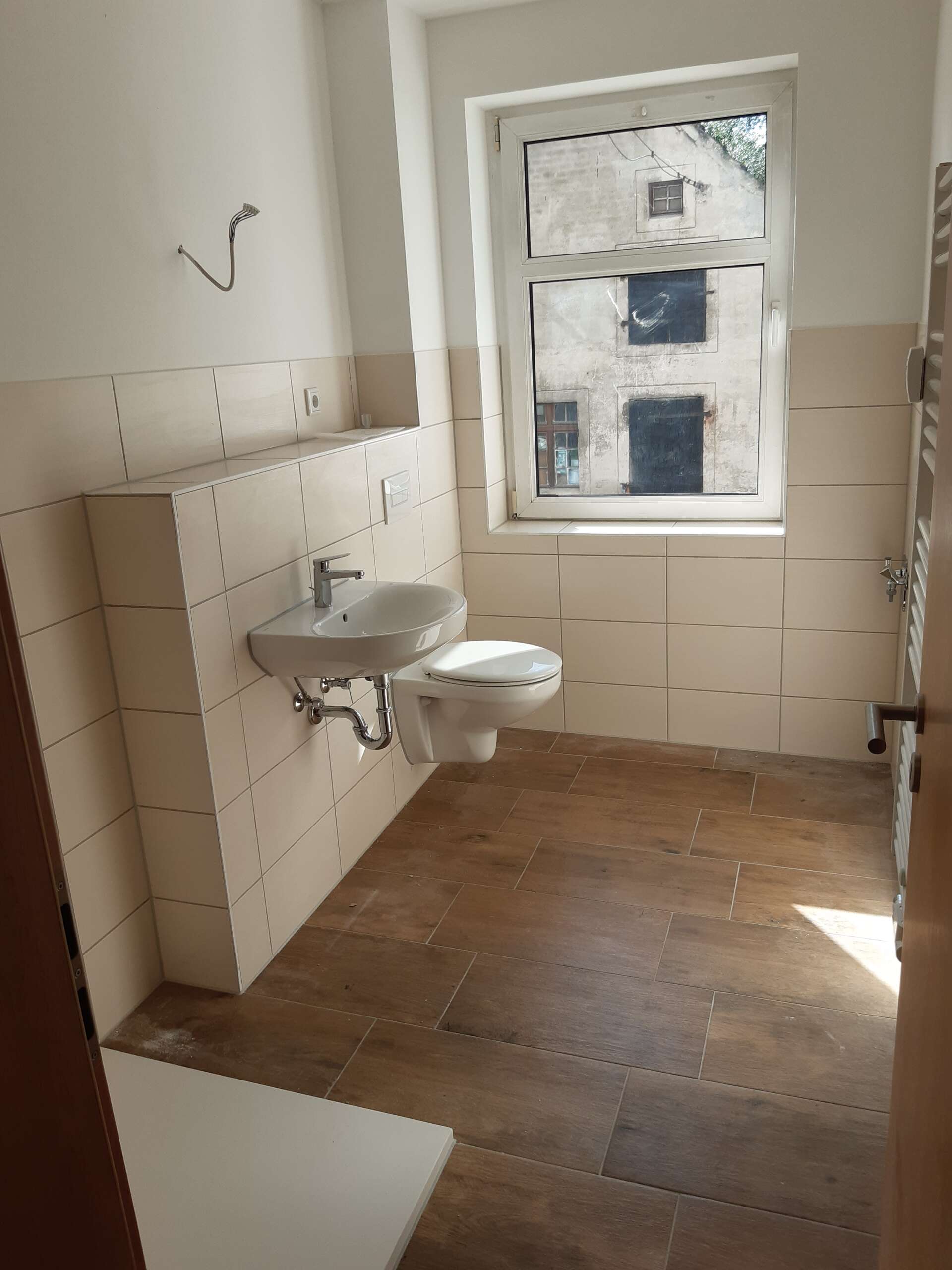 Modernes Badezimmer mit Waschbecken, Toilette und großem Fenster, Holzfliesenboden und cremefarbenen Fliesenwänden.