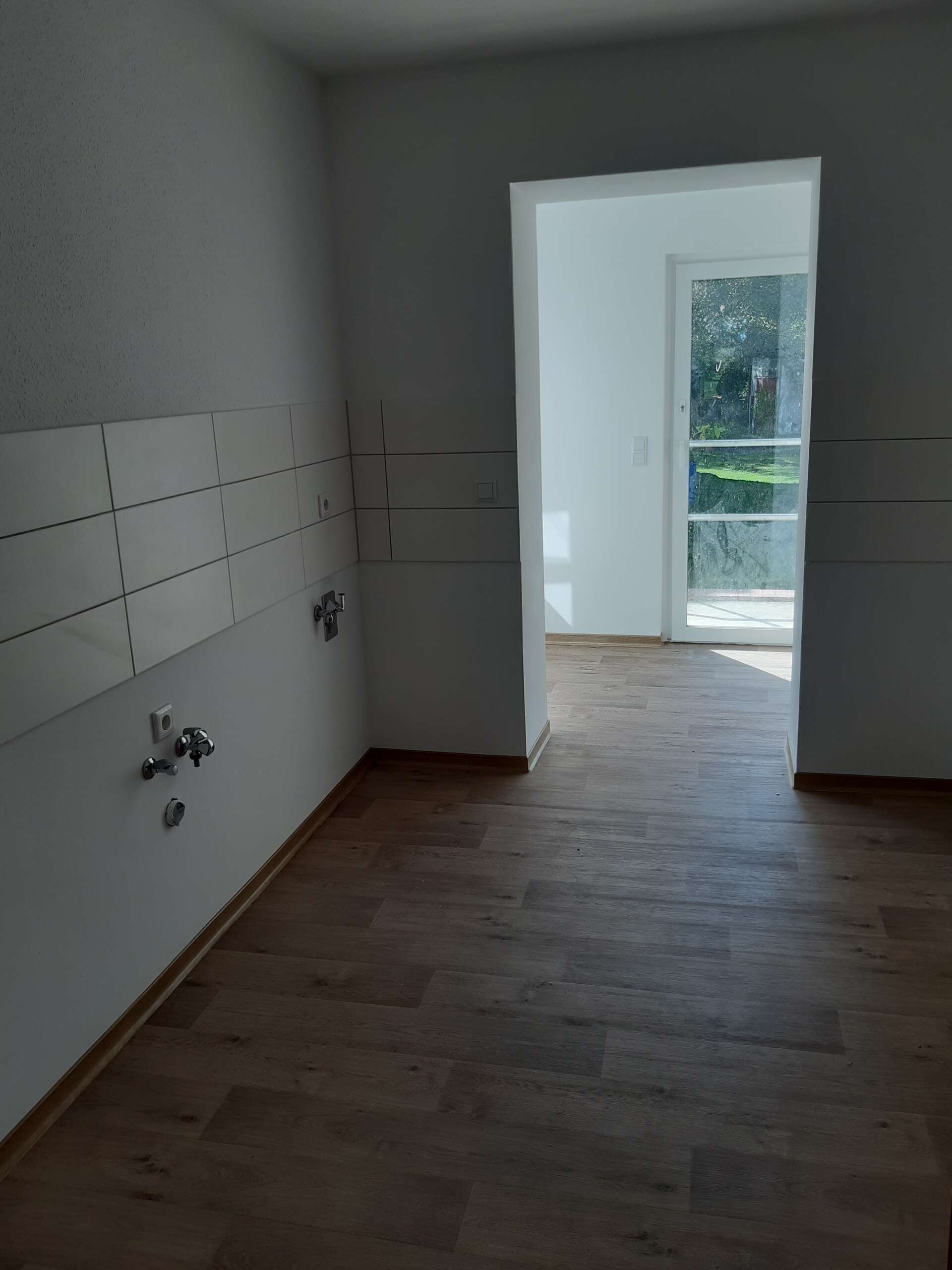Leerer Raum mit Laminatboden, Lichtdurchflutete Tür, weiße Wände, Fliesenbereich an der linken Wand, moderner Neubau.