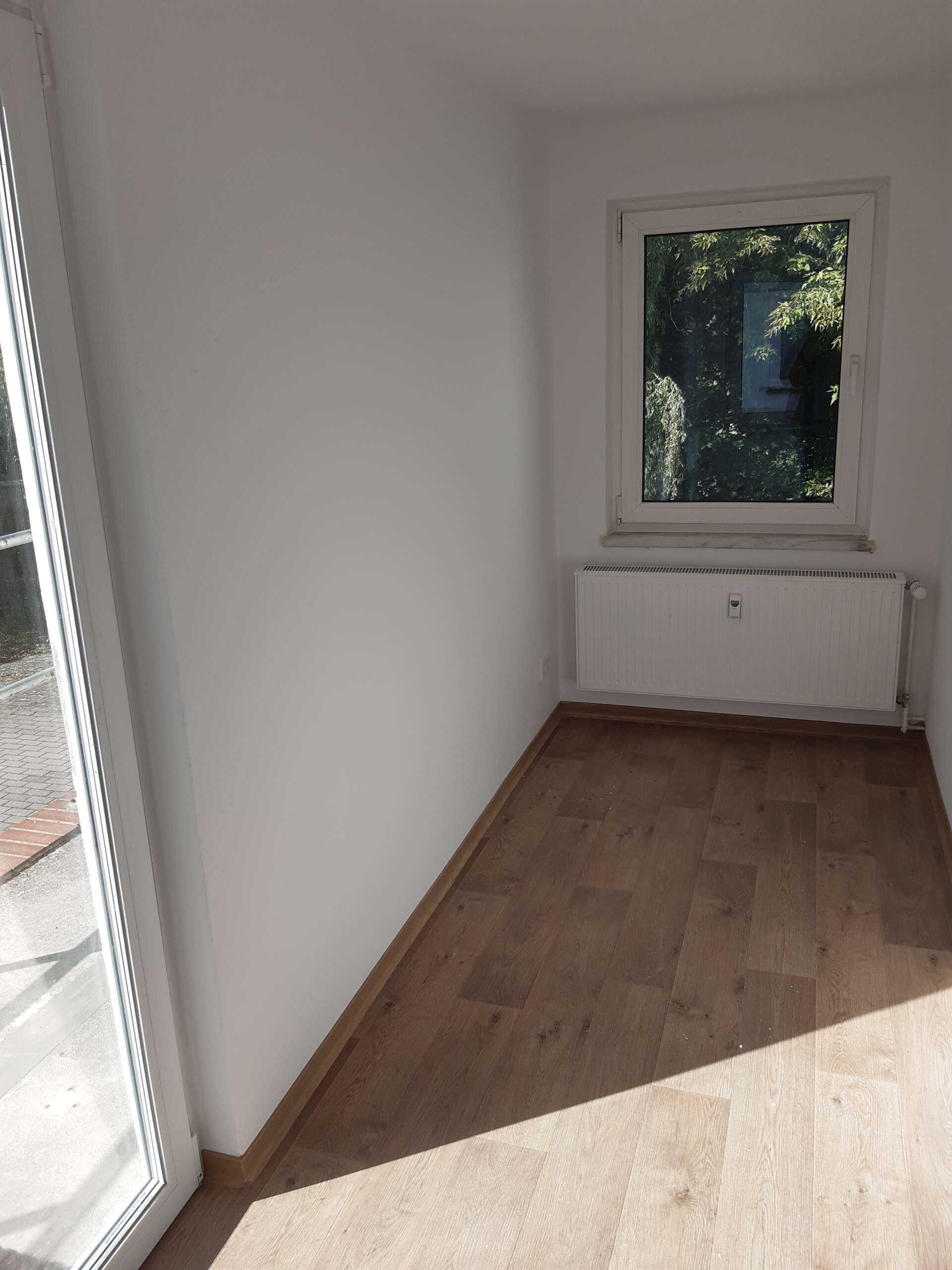 Leerer Raum mit weißen Wänden, Holzfußboden, Fenster und viel Tageslicht, Blick ins Grüne.