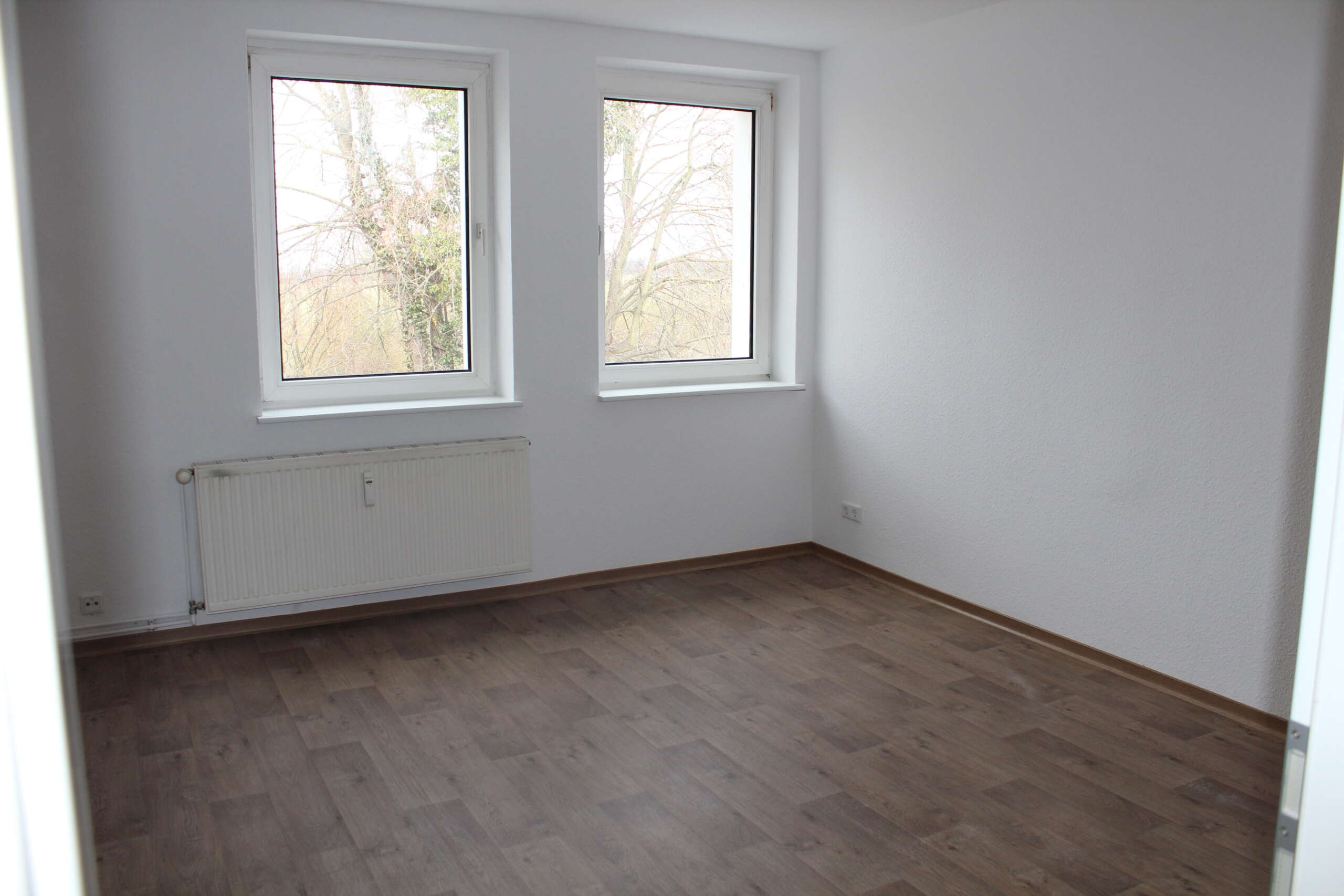 Leeres Zimmer mit Laminatboden, zwei Fenstern und Heizung, Blick auf Bäume. Weiß gestrichene Wände.