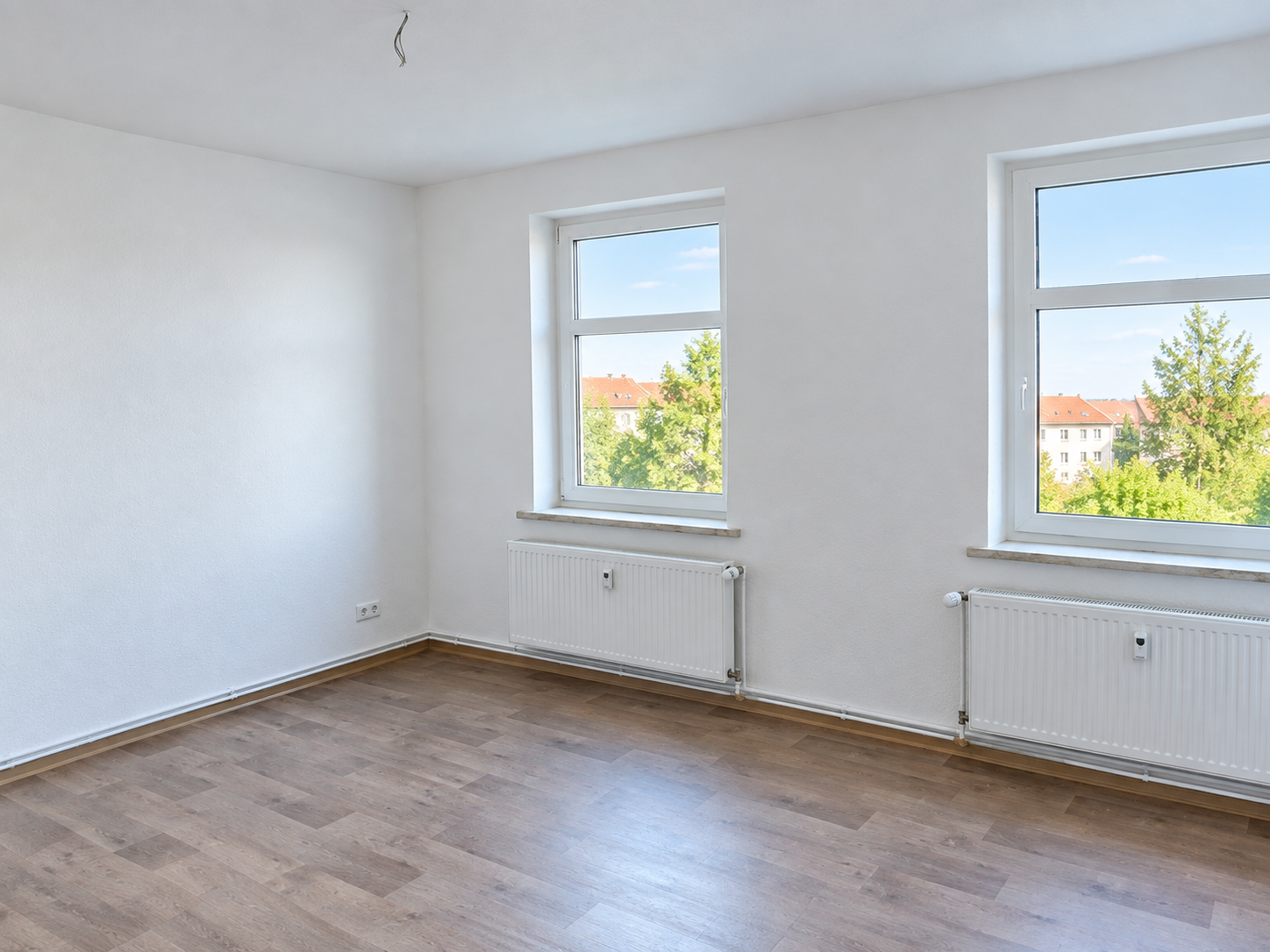 Leeres, helles Zimmer mit zwei Fenstern, weiß gestrichenen Wänden und Holzfußboden, Ausblick ins Grüne.