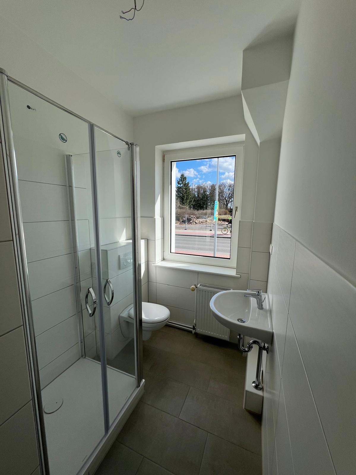 Helles Badezimmer mit Dusche, WC, Waschbecken und Fenster, das Tageslicht hineinlässt.