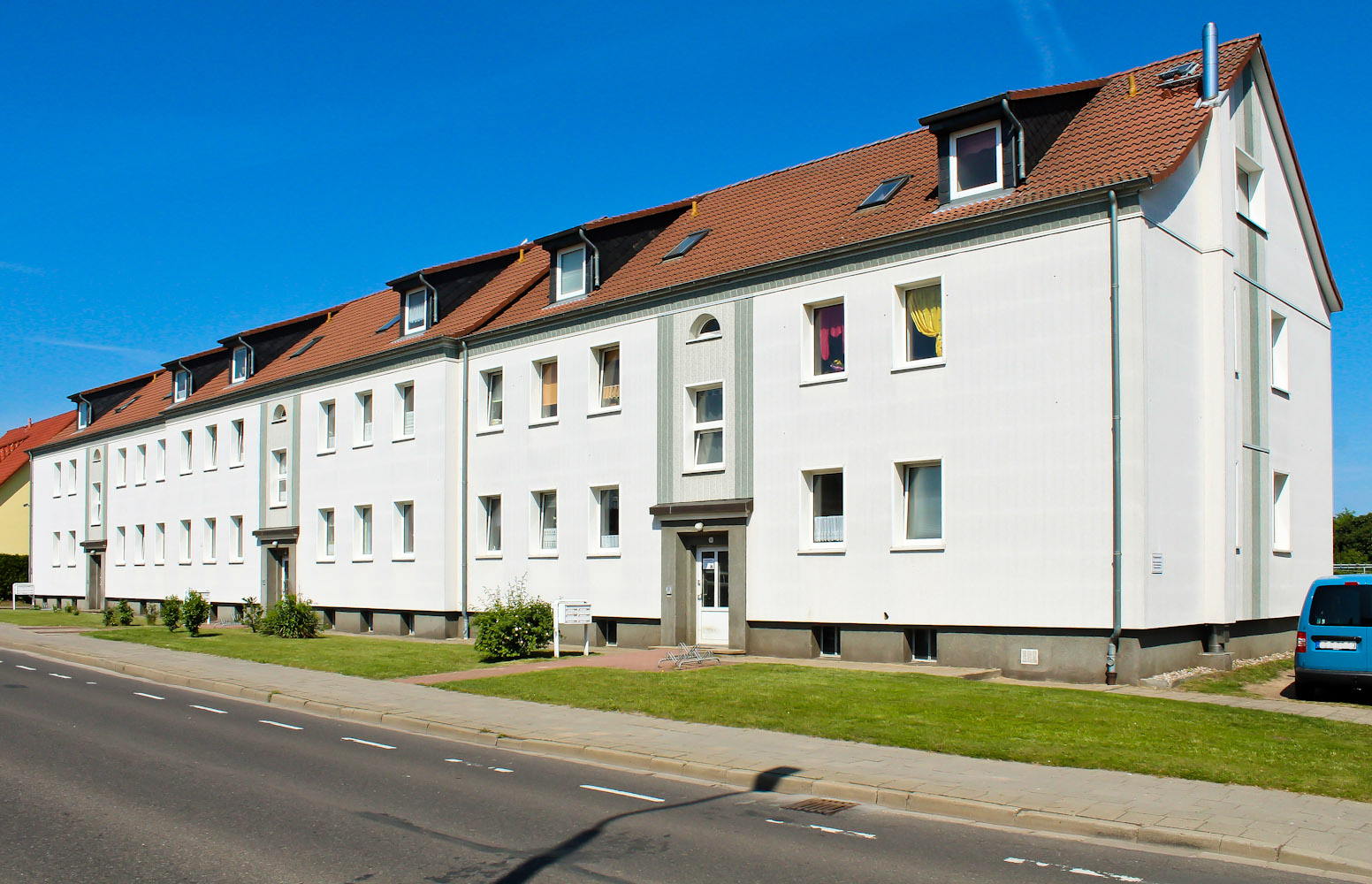 Weiße Mehrfamilienhäuser mit roten Dächern an einer Straße unter blauem Himmel.