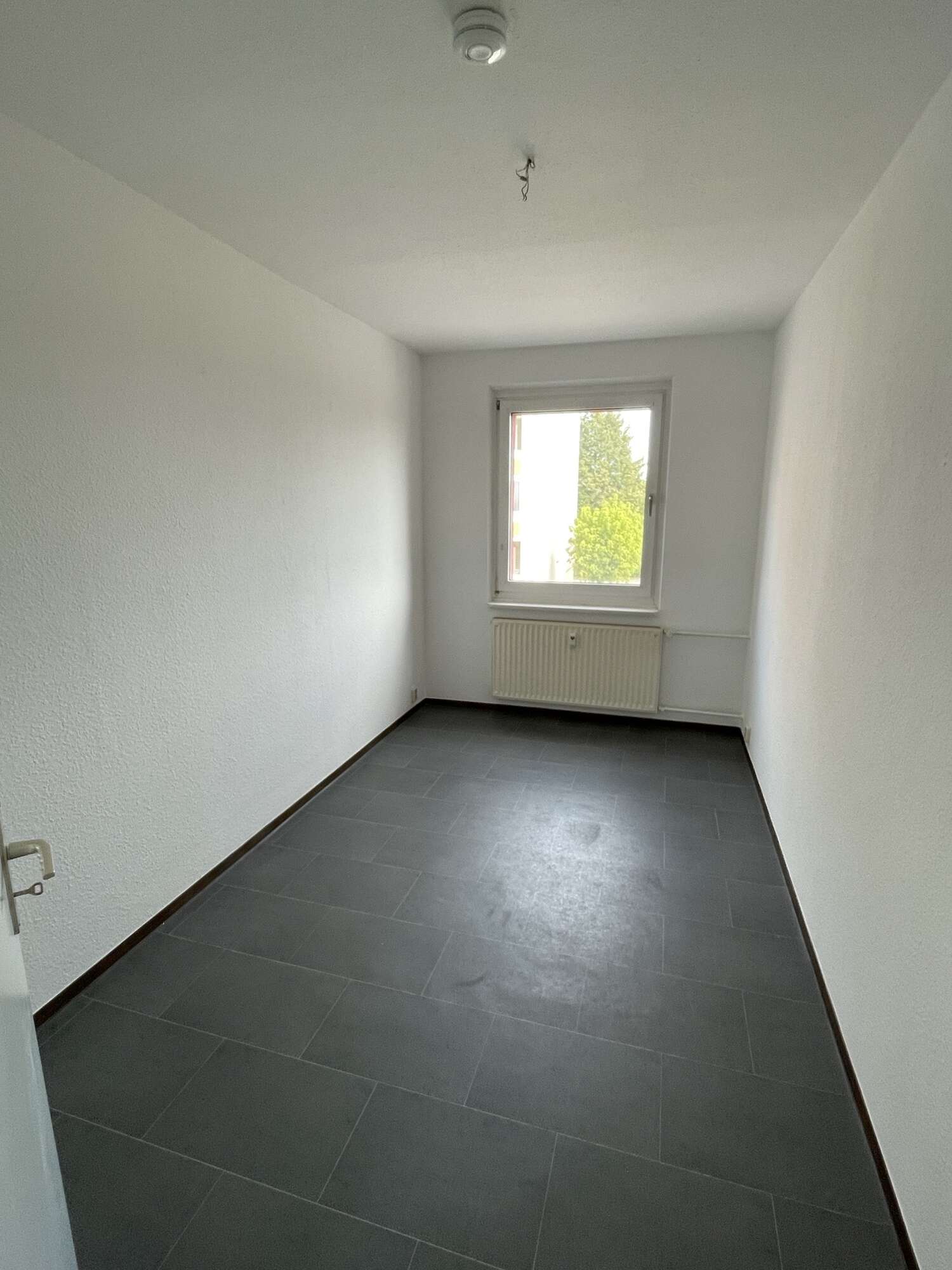 Leerer Raum mit grauem Fliesenboden, weiß gestrichenen Wänden und einem Fenster mit Blick ins Grüne.