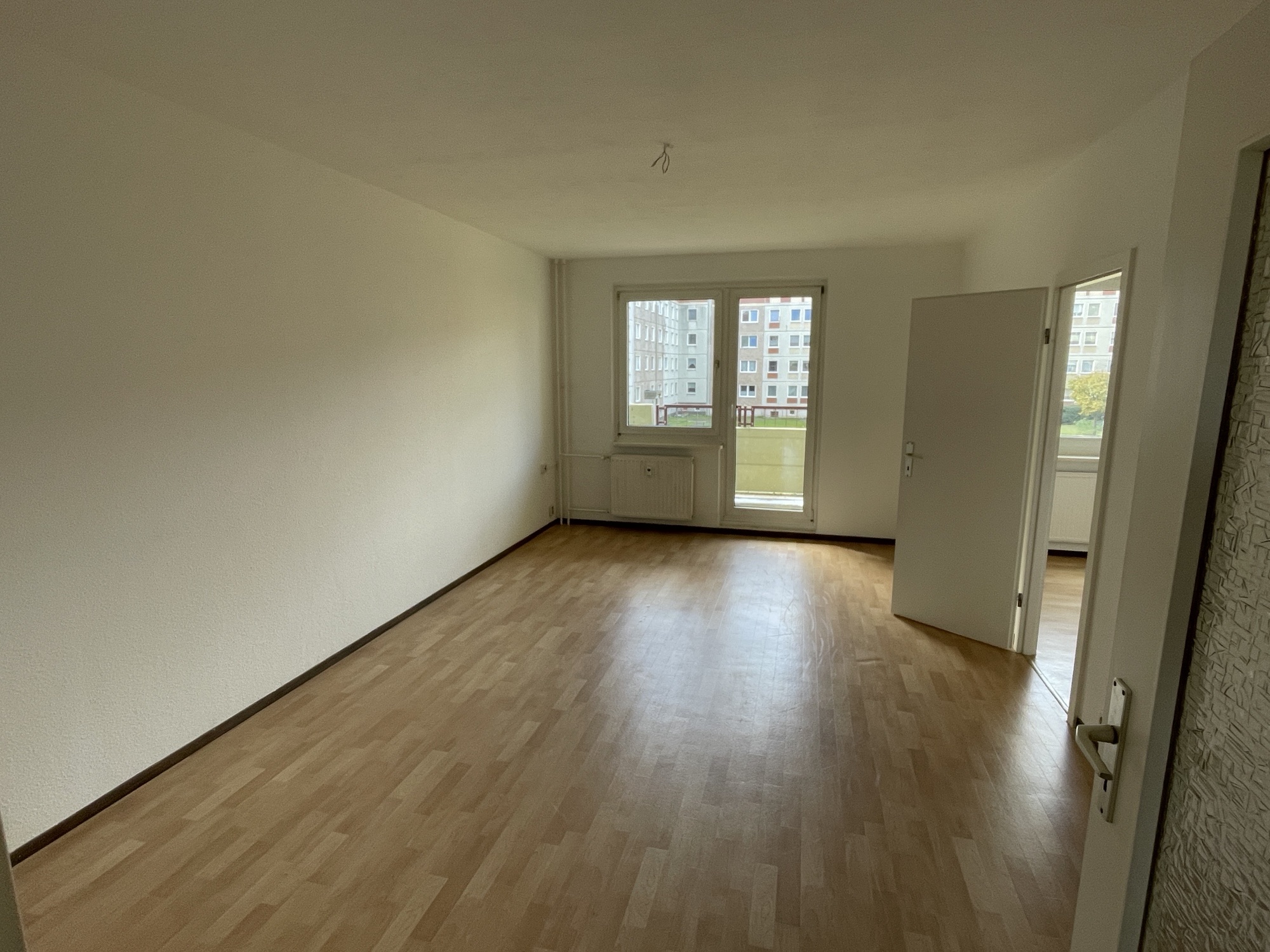 Leeres, helles Wohnzimmer mit Holzboden, großen Fenstern und offenem Zugang zu angrenzendem Raum.