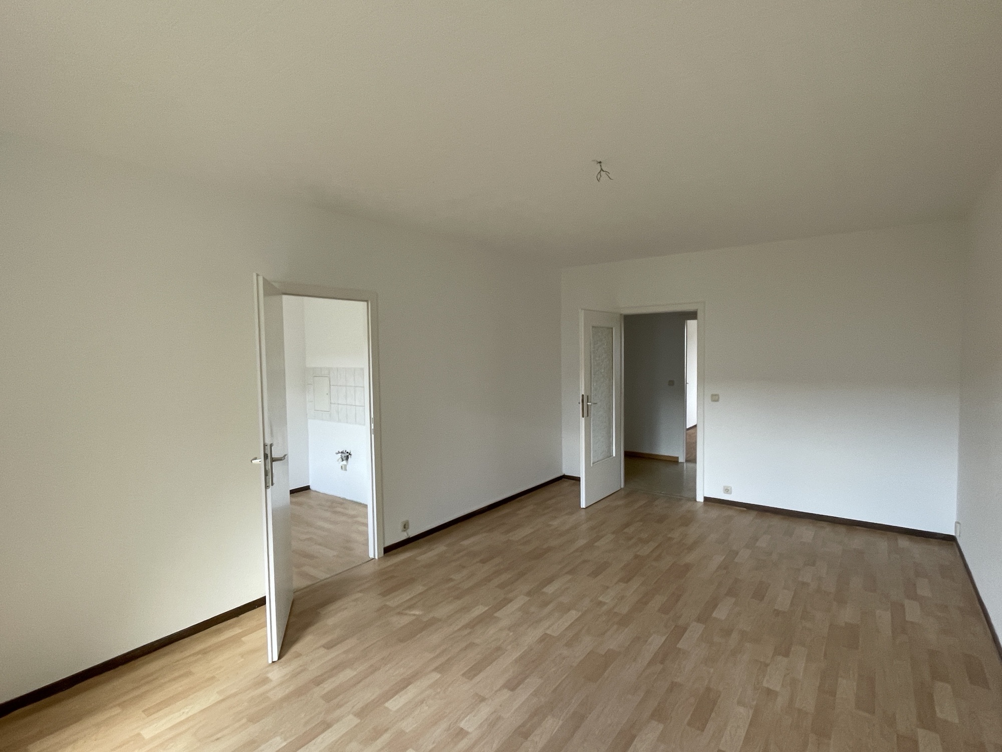 Leeres Zimmer mit Holzboden, weißen Wänden und zwei Türen, Zugang zur Küche.