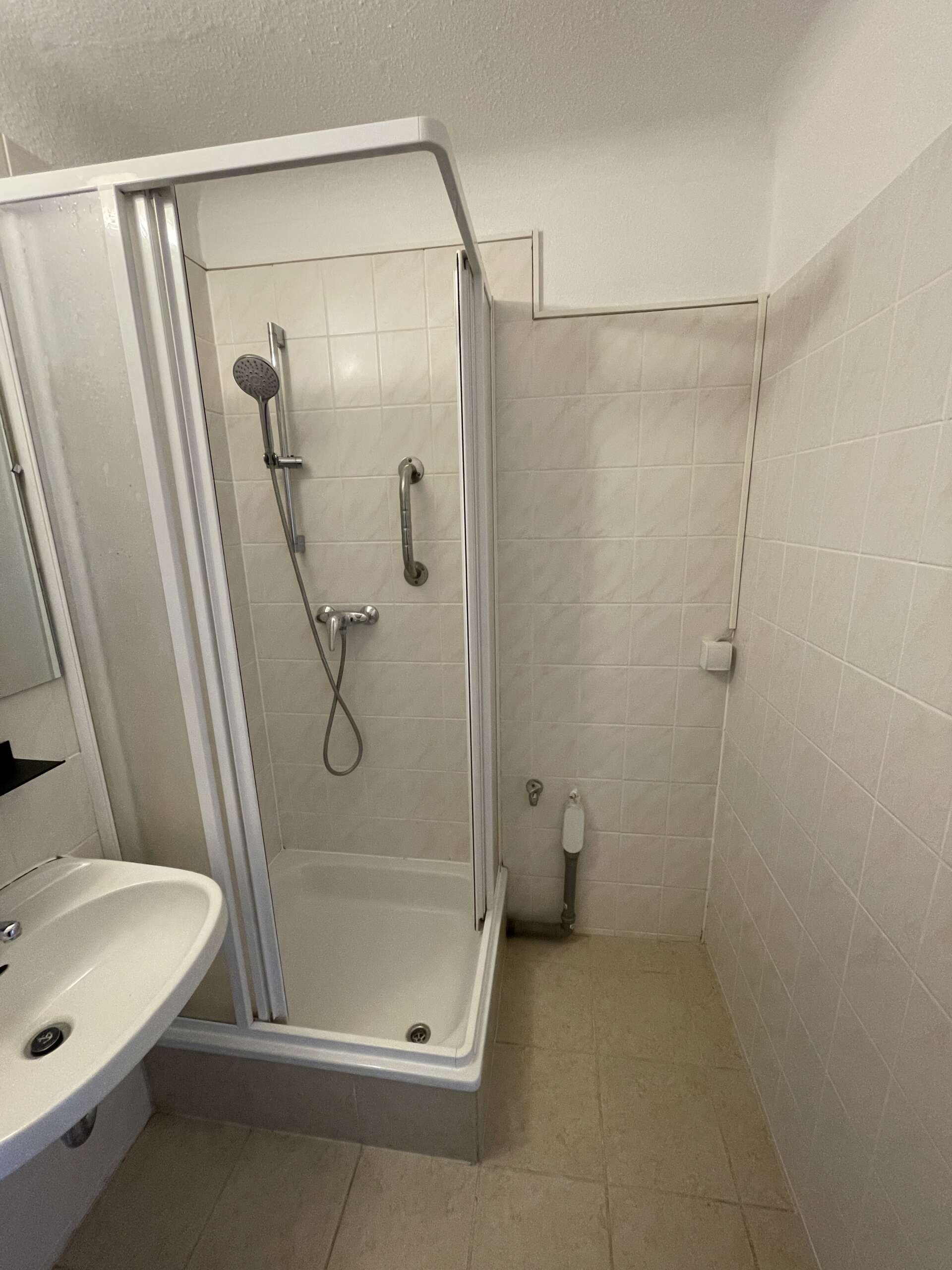 Kleine, geflieste Dusche mit Duschkopf, Griff und Waschbecken in einem beigen Badezimmer.