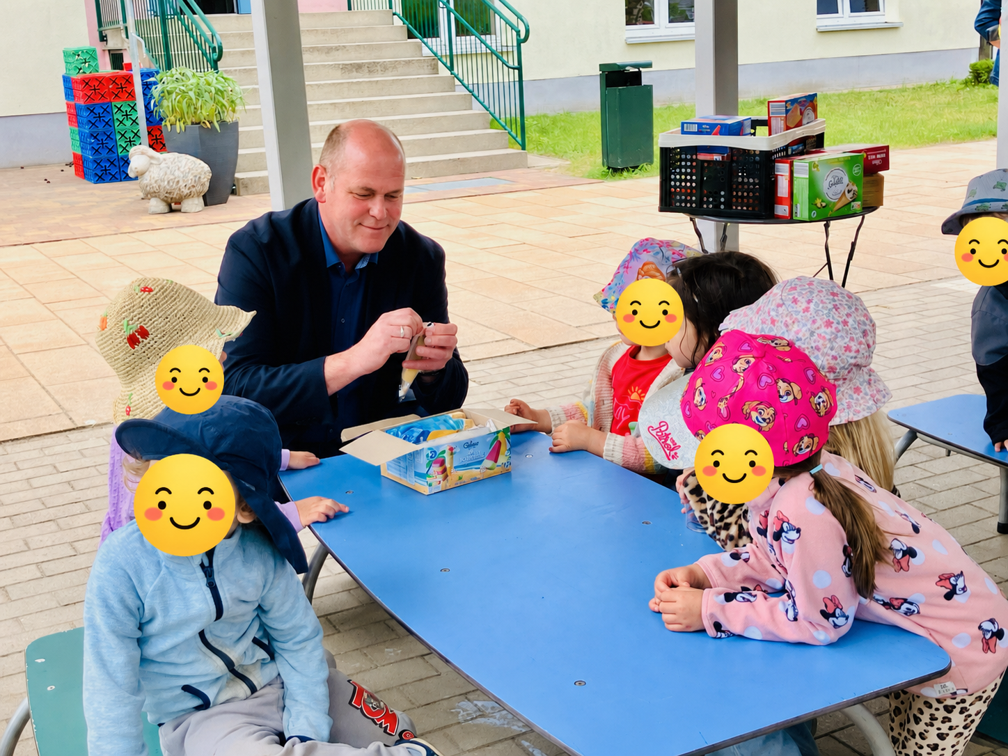 Ein Mann spielt mit Kindern im Freien an einem Tisch, bunte Hüte und Spielzeug.
