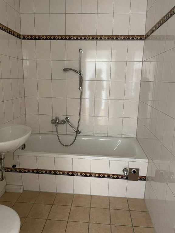 Weiß gefliestes Badezimmer mit Badewanne, Dusche, Waschbecken und Zierfliesenband auf beigem Fliesenboden.