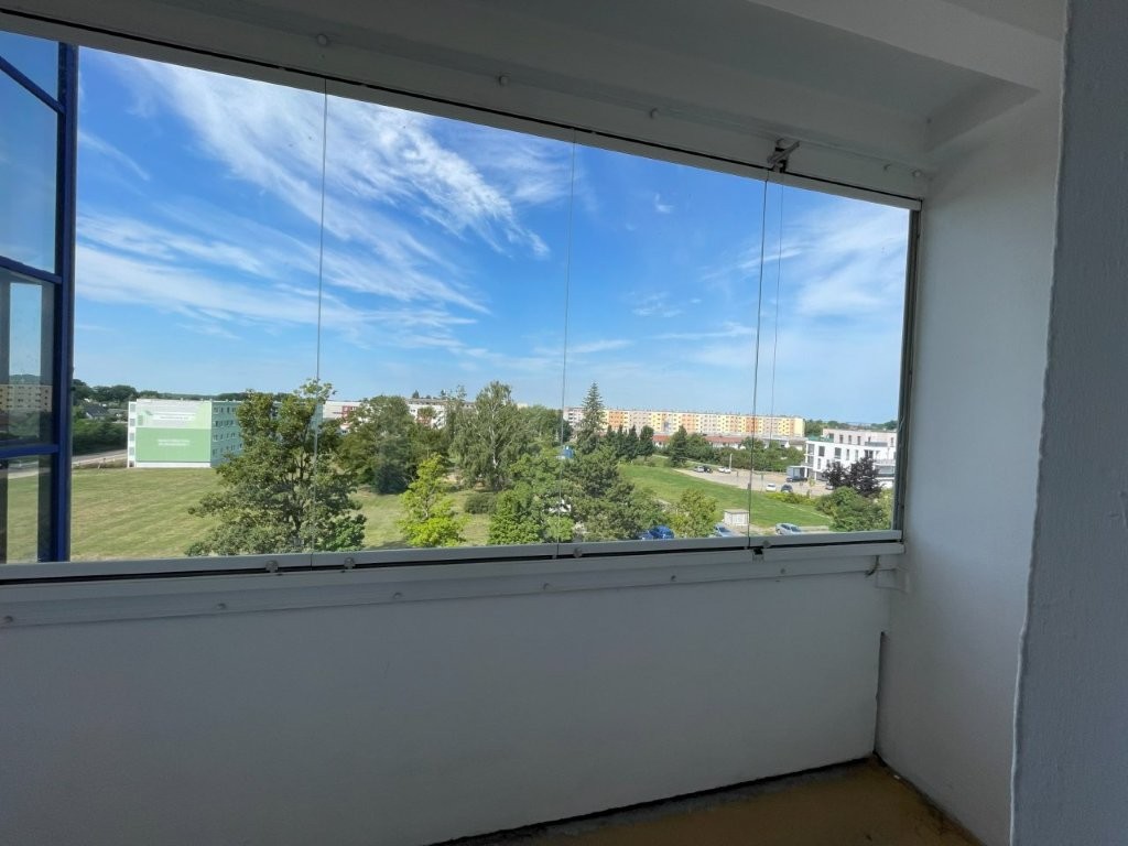 Blick von einem Balkon auf einen sonnigen Park mit Bäumen und Wohngebäuden im Hintergrund.