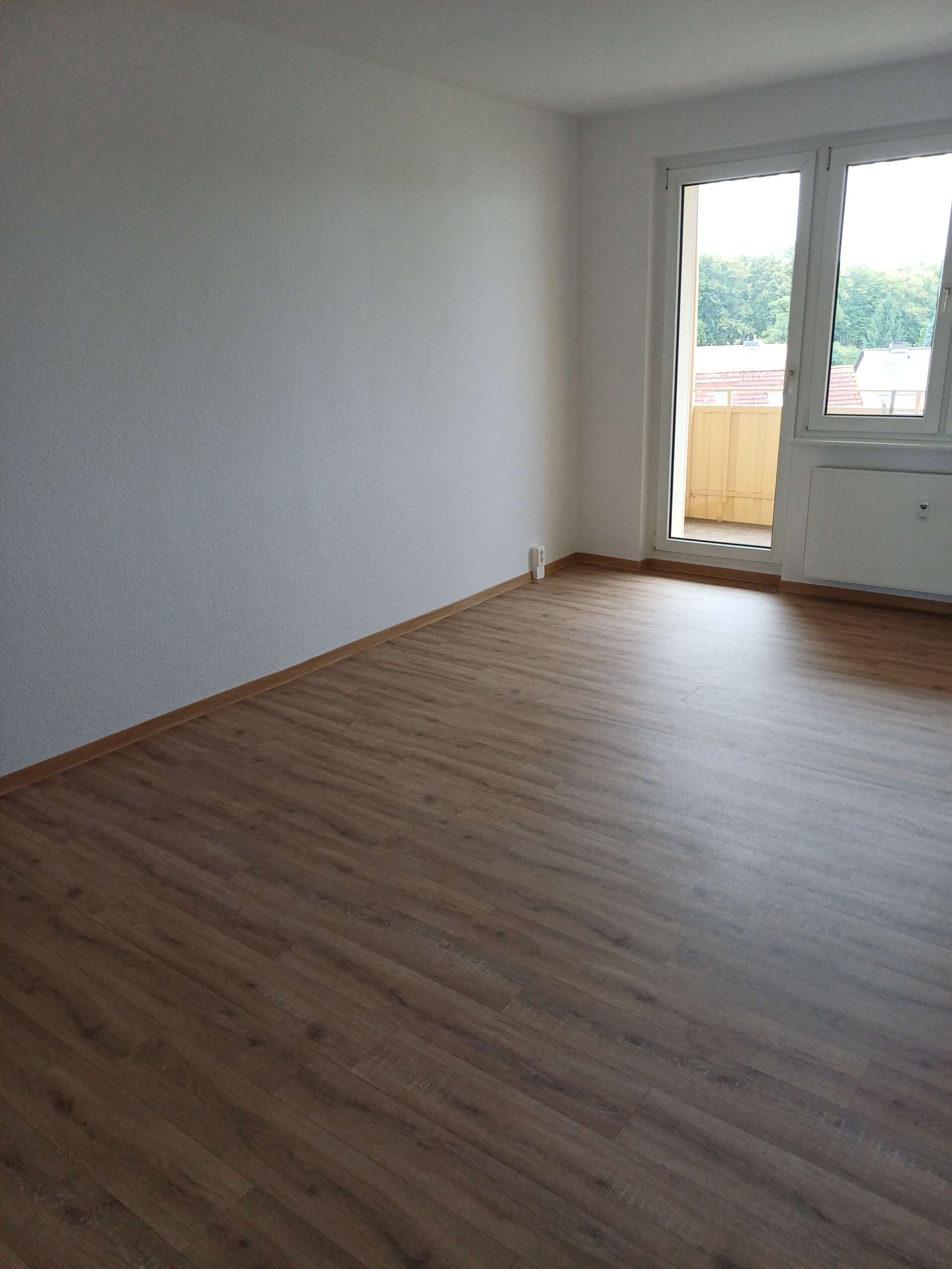 Leerer Raum mit Holzboden, weißer Wand und großen Fenstern mit Ausblick auf eine grüne Landschaft.