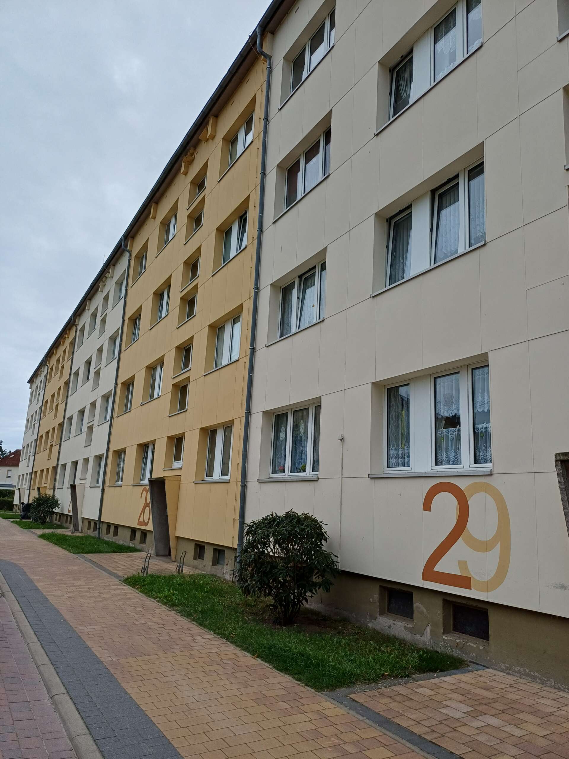 Wohnblock in beige und gelb, Nummer 29 groß auf Fassade, seitlicher Blick auf Gehweg und Grasfläche.