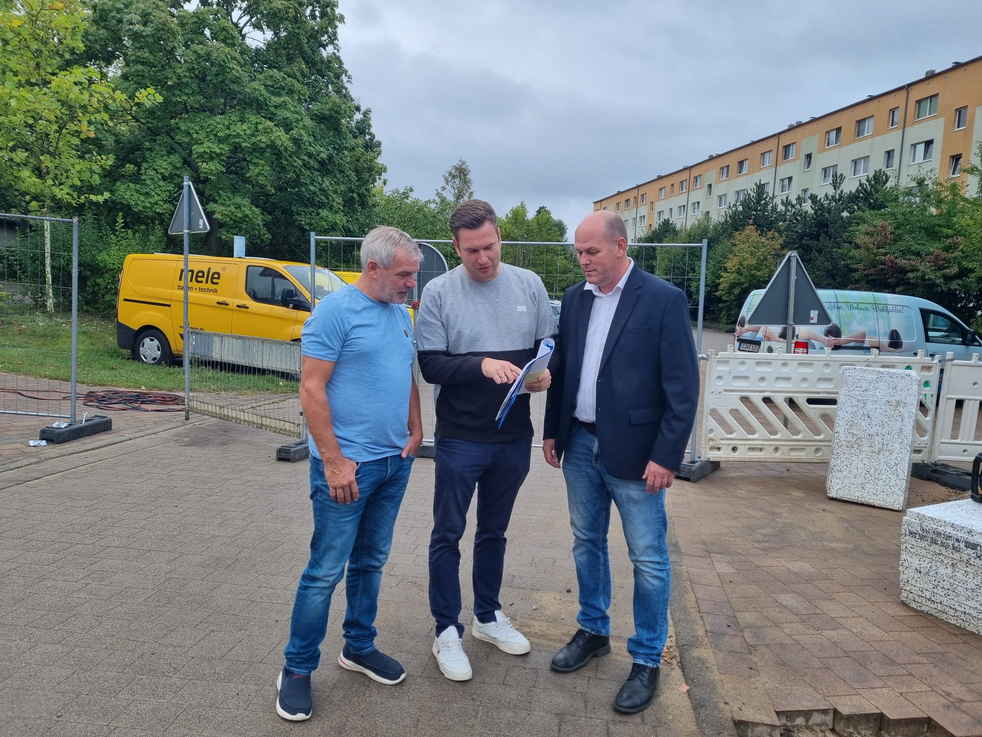 Drei Männer diskutieren Baupläne auf einer Baustelle mit gelbem Lieferwagen im Hintergrund.